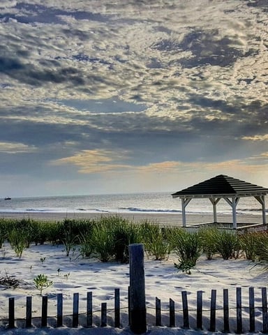 Beach at sunset with wooden gazebo, dune fence, beach grass, and ocean horizon under dramatic cloudy sky