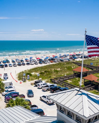 American flag flying over crowded beach parking lot with ocean, sand dunes, and beachgoers visible