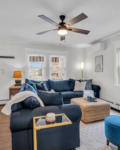Bright living room with dark blue sectional sofa, woven ottoman, blue accent chairs, ceiling fan, and hardwood floors
