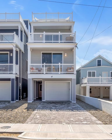 Two modern three-story beach houses with white railings, multiple decks, and ground-level garages facing a paved driveway.