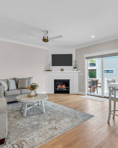 Open-plan living room with fireplace, TV, gray seating, hardwood floors, and kitchen with stainless steel appliances visible on right