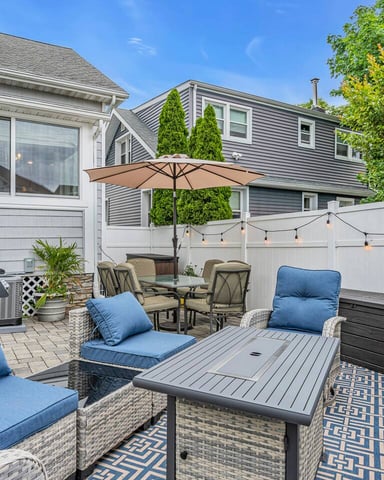 Backyard patio with wicker seating, blue cushions, fire table, umbrella, string lights, and white privacy fence