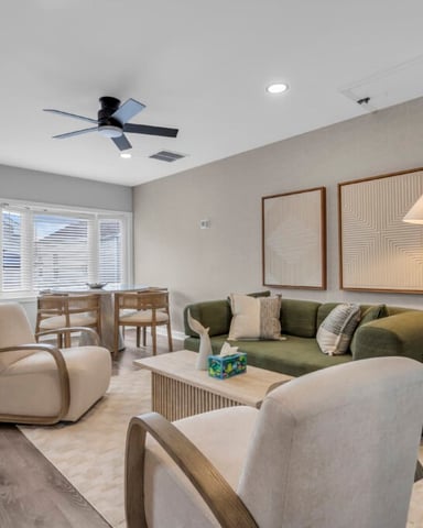 Open-plan living room with beige armchairs, green sofa, ceiling fan, and dining area visible through windows