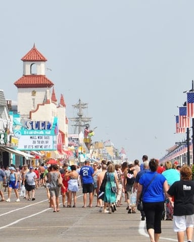 Crowded boardwalk with shops, castle-themed building, American flags, and clear sky
