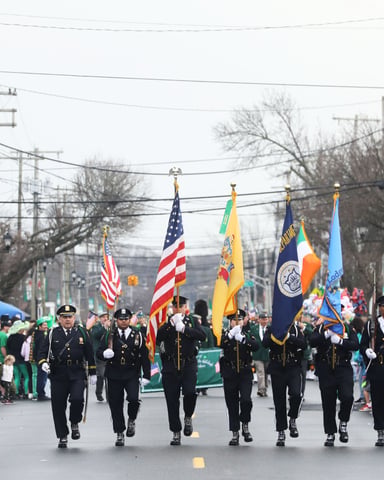 St. Patrick's Day parade with uniformed military personnel carrying American and state flags down a street lined with spectators
