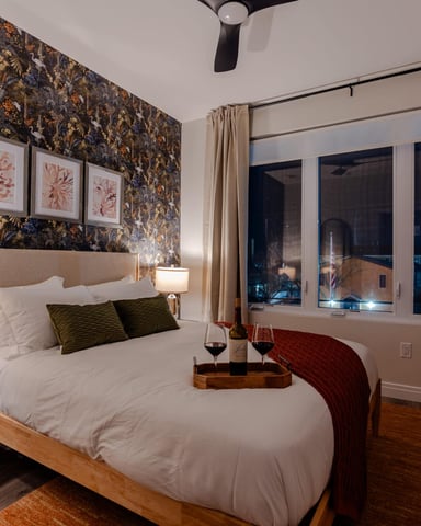 Bedroom with patterned wallpaper, wood bed frame, white bedding, framed coral artwork, large windows with city night view, and beige curtains.