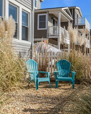 Two turquoise Adirondack chairs on gravel patio surrounded by dried ornamental grasses, with beach house and dunes in background