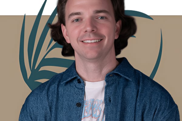 Man with shoulder-length dark hair wearing denim shirt over graphic tee, smiling at camera against tan background with teal plant illustration