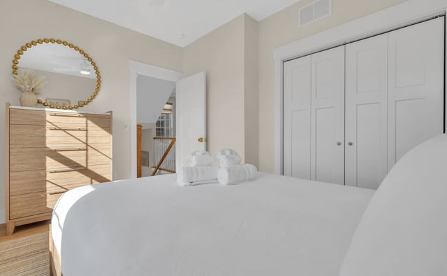 Bright bedroom with white bed, wood dresser, gold-framed mirror, white closets, and staircase visible through doorway