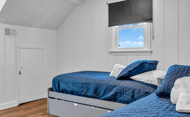Bright attic bedroom with two twin beds, blue quilts, white pillows, window with black shade, and wooden floor