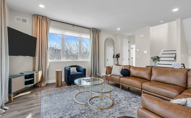 Modern living room with tan leather sofa, blue armchair, glass coffee table, wall-mounted TV, and large windows overlooking bare trees.
