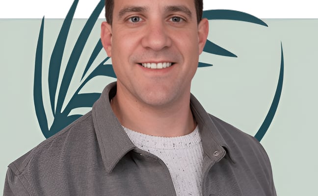 Man smiling at camera wearing gray shirt over white sweater, tropical leaf background