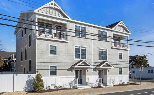 Modern white shingle house with dual peaked roofs, second-floor balconies, and two front entrances under a clear blue sky.