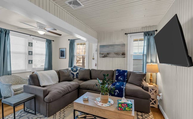Bright living room with gray sectional sofa, wood coffee table, wall-mounted TV, and teal curtains framing multiple windows with ocean view.